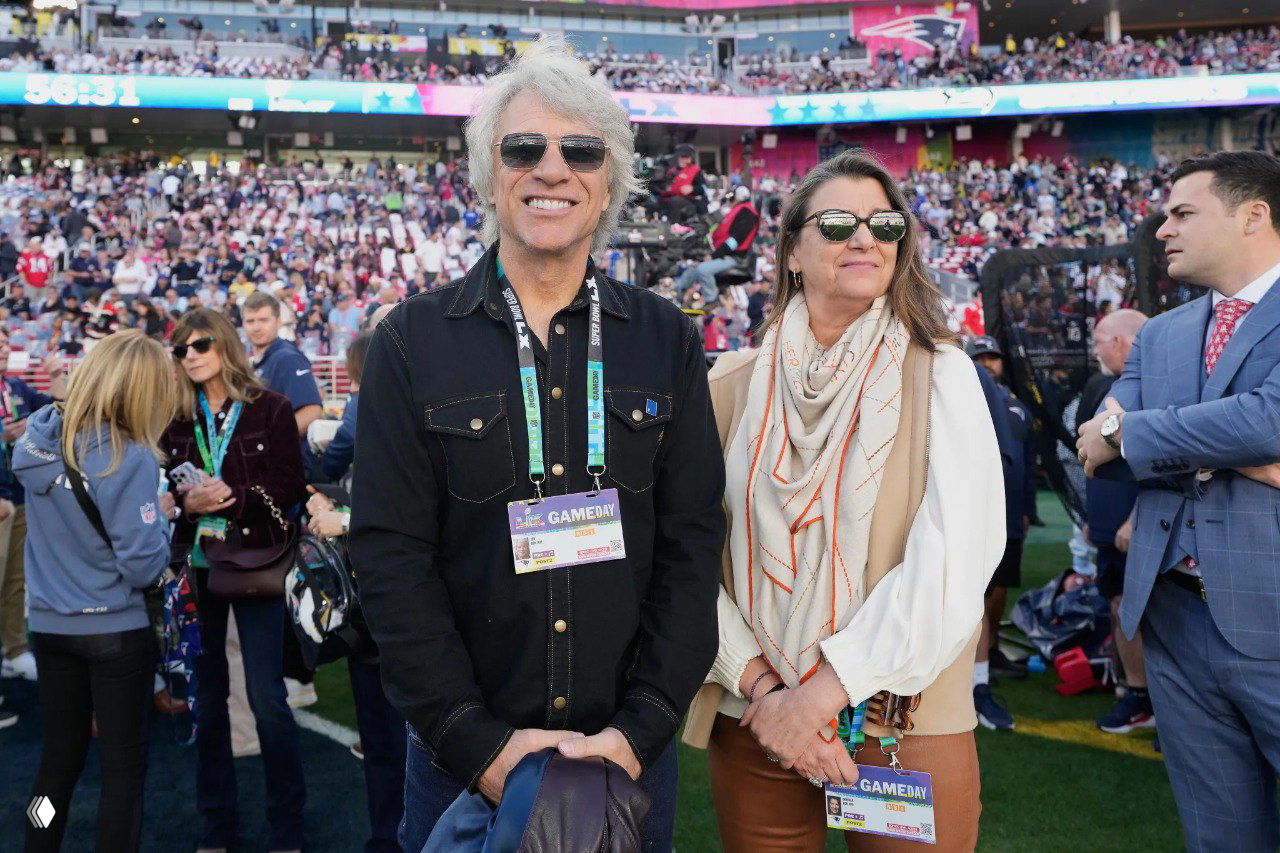 Пара гостей улыбается на поле стадиона: мужчина с седыми волосами и женщина рядом, на заднем плане видна толпа зрителей и атмосфера матча