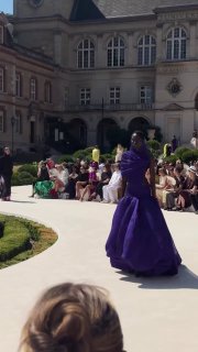 Показ Balenciaga на кампусе Cité Universitaire: модель в фиолетовом объёмном платье идёт по дорожке на фоне зрителей и архитектуры.