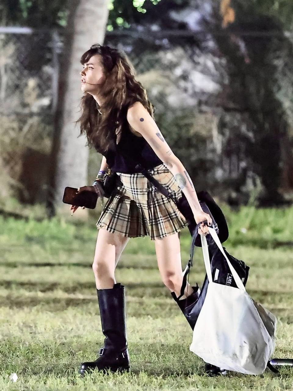 Девушка в мини‑юбке в клетку и высоких сапогах несёт сумки и вещи, идёт по травяному полю в ночное время фестиваля — гранж‑эстетика в образе.