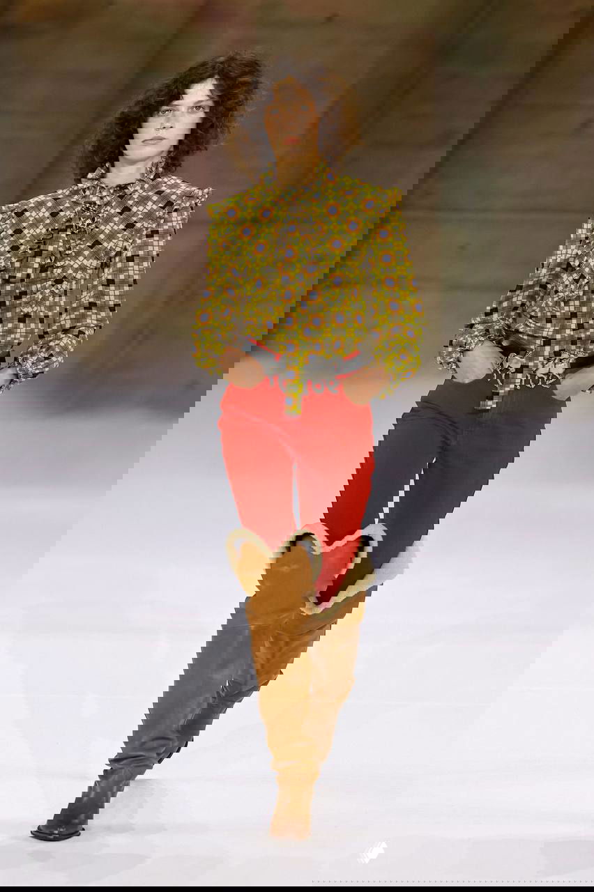 Модель в клетчатой рубашке и высоких сапогах с меховой отделкой шагает по дымчатому подиуму — ковбойские мотивы и загородная эстетика в осеннем ключе.