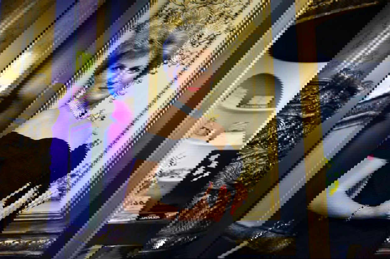 Восковая фигура принцессы Дианы в черном платье на фоне золотистого декора и колонн музея; в кадре видны элементы оформления экспозиции и техника.