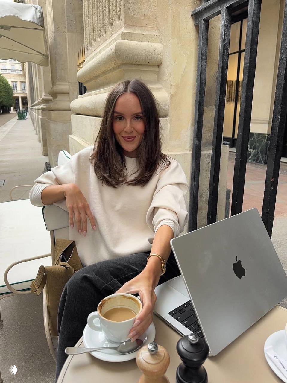 Девушка в белом свитере за столиком в кафе с чашкой кофе и открытым ноутбуком, атмосферный кадр рабочего перерыва на улице.