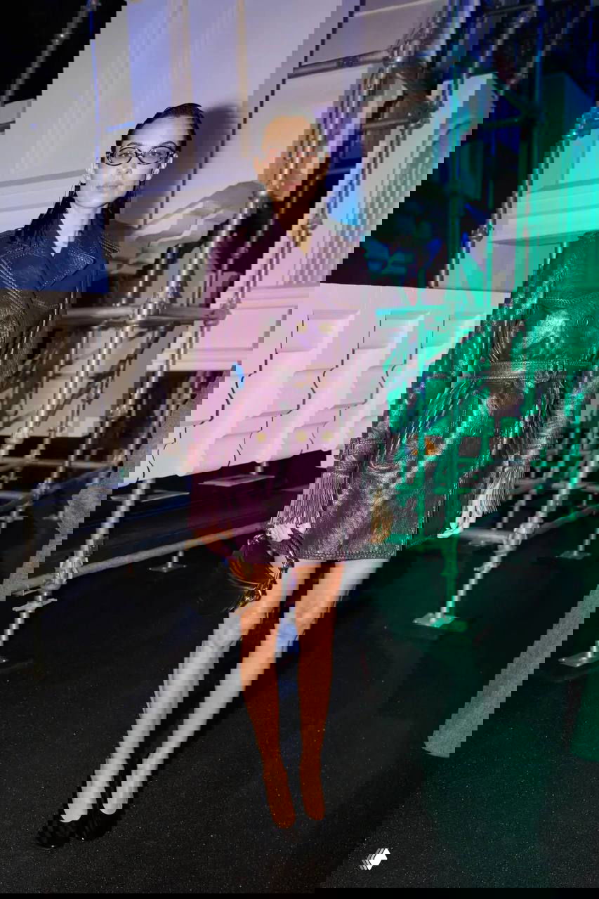 Гостья в фиолетовом кожаном тренче и очках стоит в интерьере шоу — кожаный плащ и минималистичный макияж в акцентном свете.