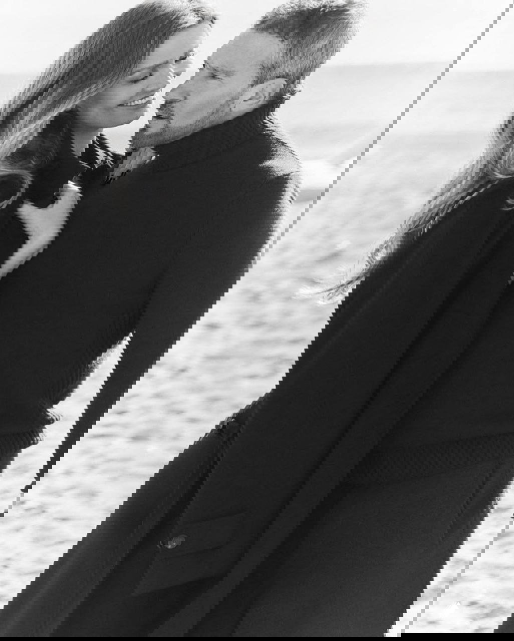 Крупный чёрно‑белый портрет: Николь Кимпел и Антонио Бандерас в пальто и свитере, обмениваются нежными взглядами на побережье.