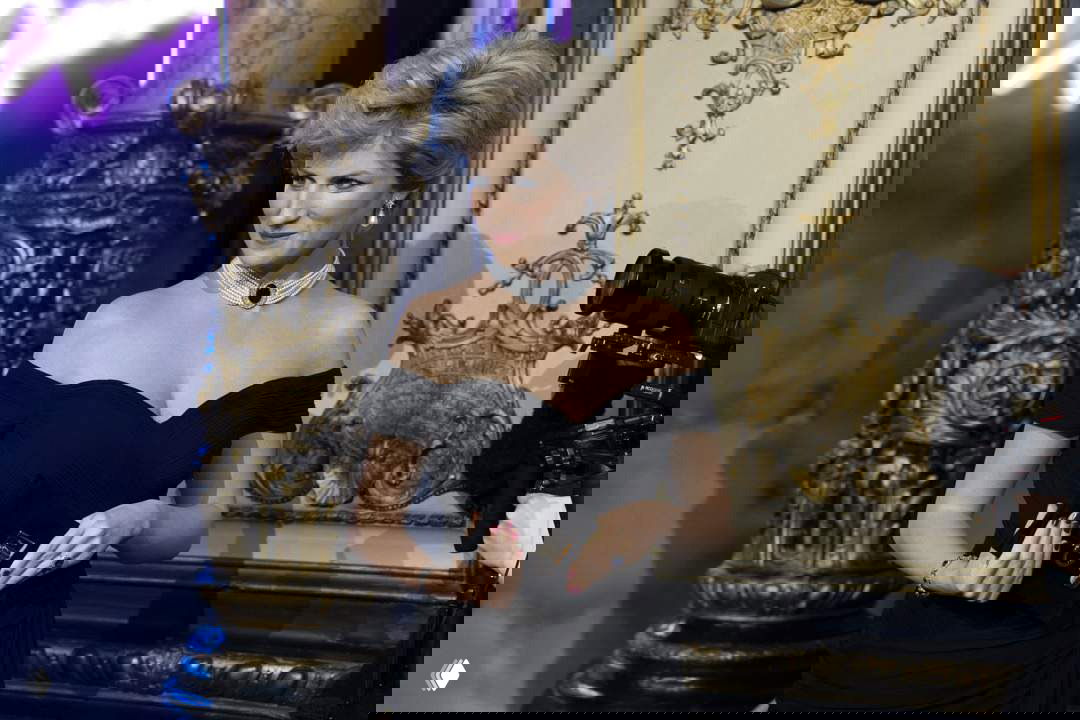 Близкий план восковой фигуры Дианы в черном платье: видны черты лица, украшения и присутствие фотографов у подиума во время презентации.