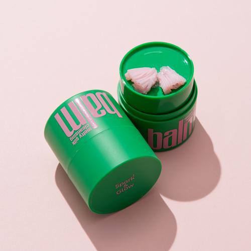 Открытая зелёная баночка бальзама Spark'&Glow Melty silk с розовыми кусочками продукта на пастельном фоне.