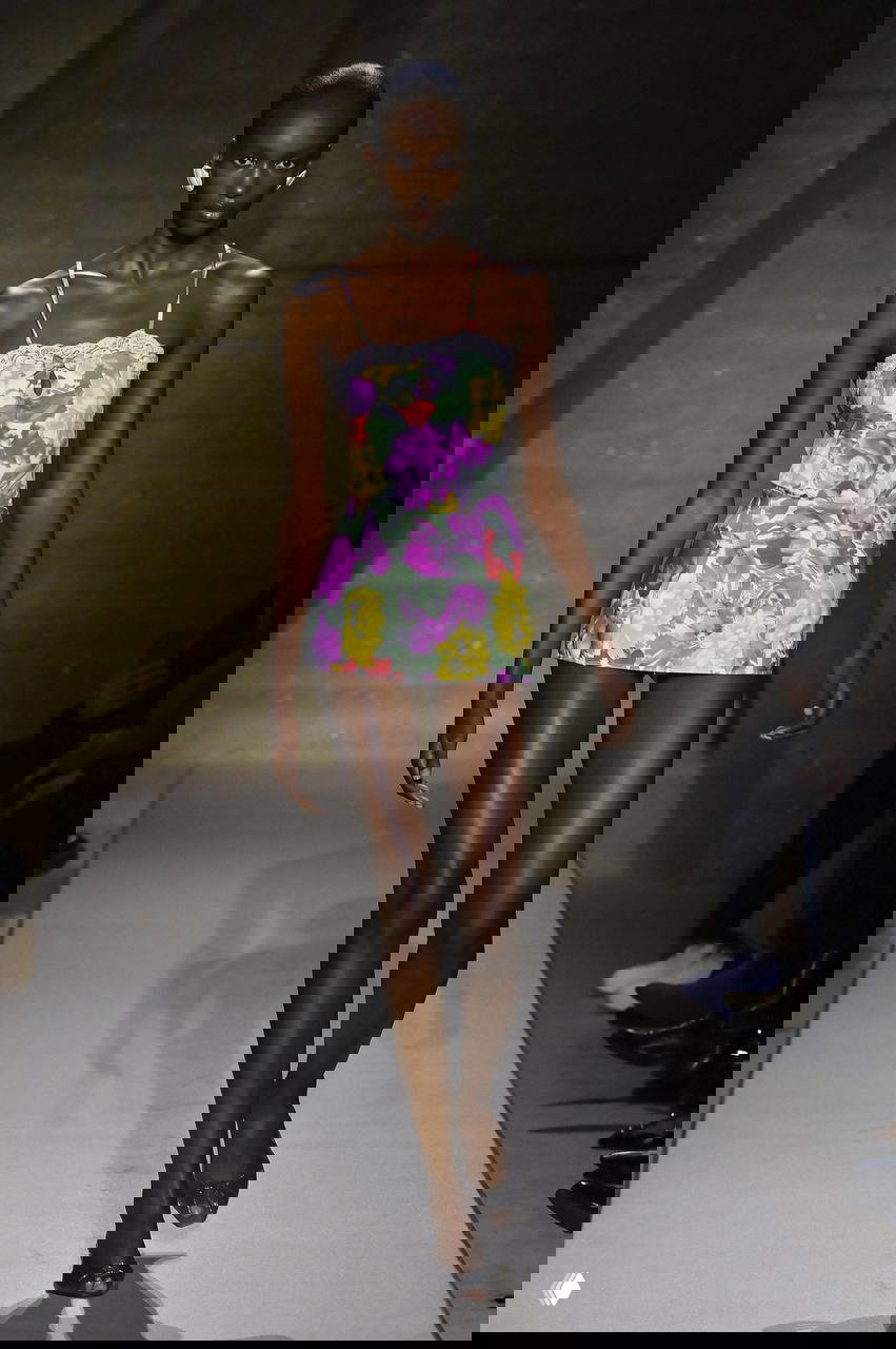 Модель в коротком цветочном платье на тонких бретелях идёт по подиуму; лёгкий летний силуэт с ярким принтом и минимальными аксессуарами.