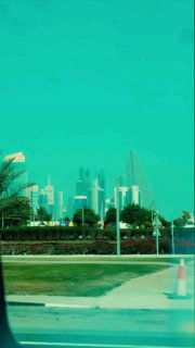 Панорамный вид на набережную и небоскрёбы Дохи через стекло автомобиля; зелёная зона и современная архитектура на фоне бирюзового неба.