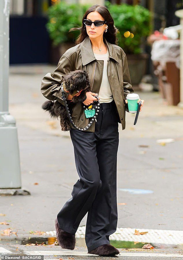 Женщина в оливковой кожаной куртке и широких брюках переходит улицу Нью-Йорка, несёт маленькую тёмную собаку и стакан в руках.