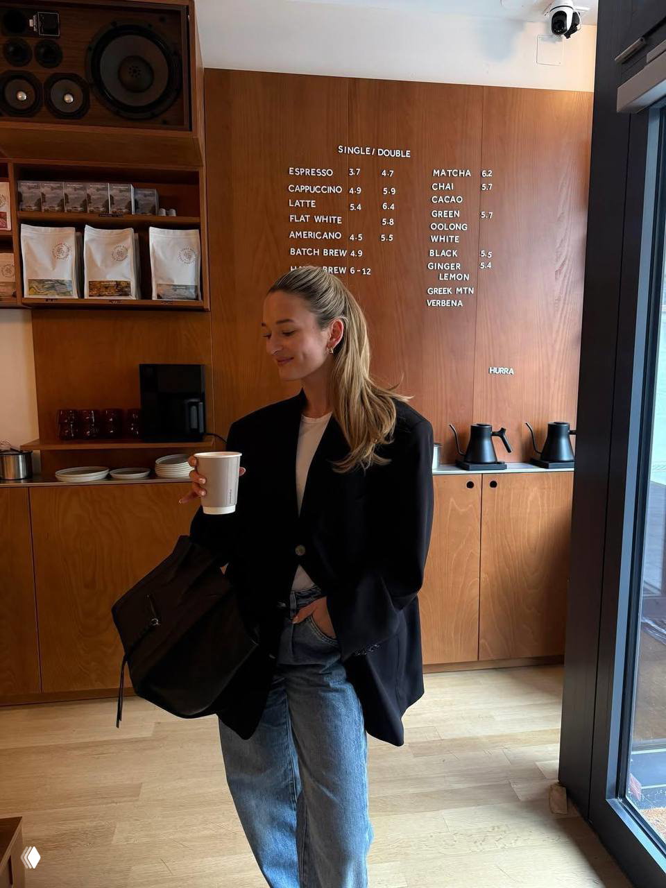 Интерьер кафе с меню на стене и девушка в чёрном пиджаке держит бумажный стакан с напитком, уютный кадр рабочего утра.