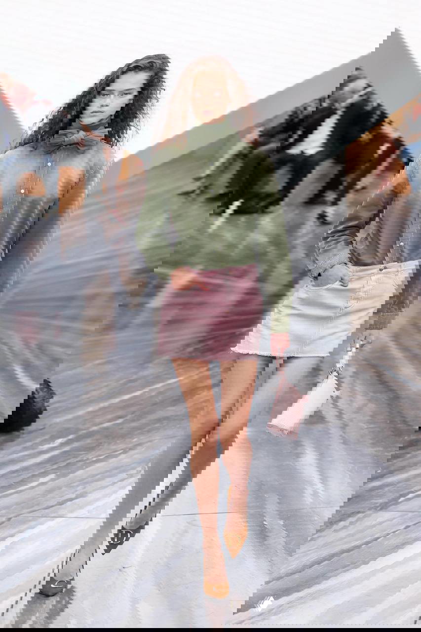 Модель в короткой розовой мини-юбке и зелёной рубашке с бантом на глянцевом подиуме, демонстрация тренда экстра‑мини и аксессуаров лета 2026.