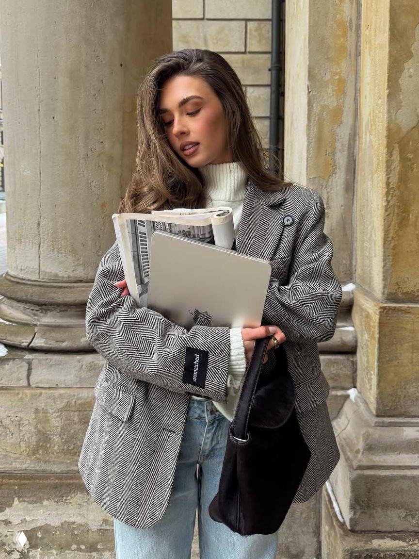 Женщина у колонн держит ноутбук и газету в пиджаке и джинсах; модный образ сочетания работы и личного стиля на улице.