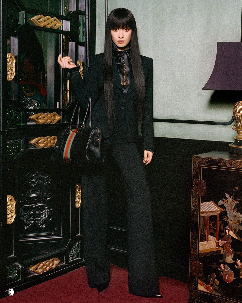 Модель в чёрном костюме и сумкой Gucci позирует на фоне тёмной резной мебели и зелёной стены в рамках кампейна The Gathering от Gucci.