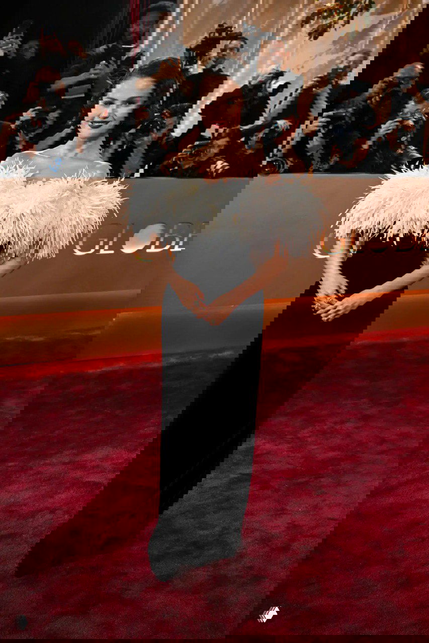 Женщина в черном платье с объемной отделкой из перьев у линии фотографов на красной дорожке — вечерний образ и крупный план.