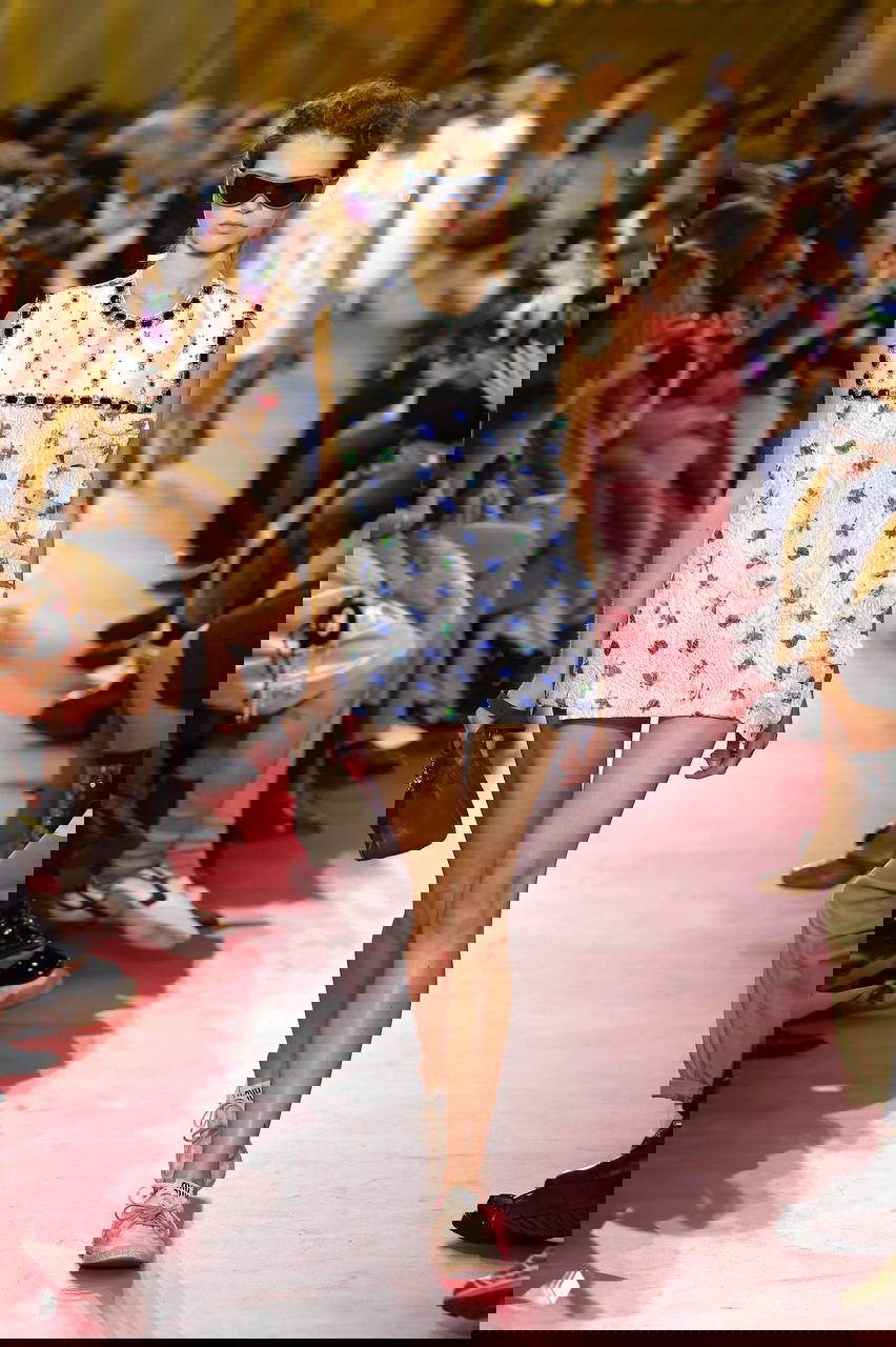 Модель в коротком платье-футляре с узором и солнцезащитными очками, дефиле на ярком розовом подиуме — акцент на мини и ретро‑эстетику.