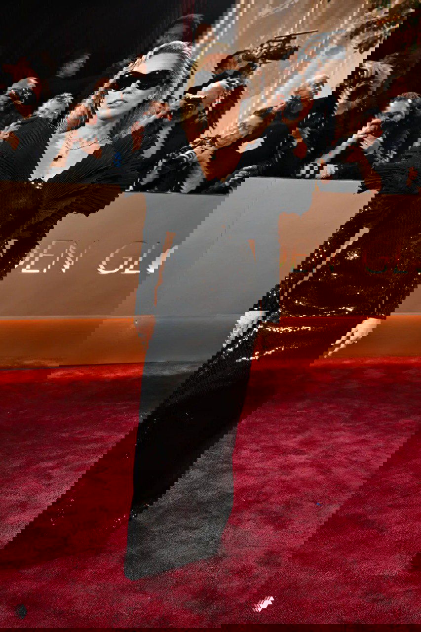 Фигура в блестящем черном платье с выразительными рукавами и затемнёнными очками на фоне папарацци и декора мероприятия на красной дорожке.