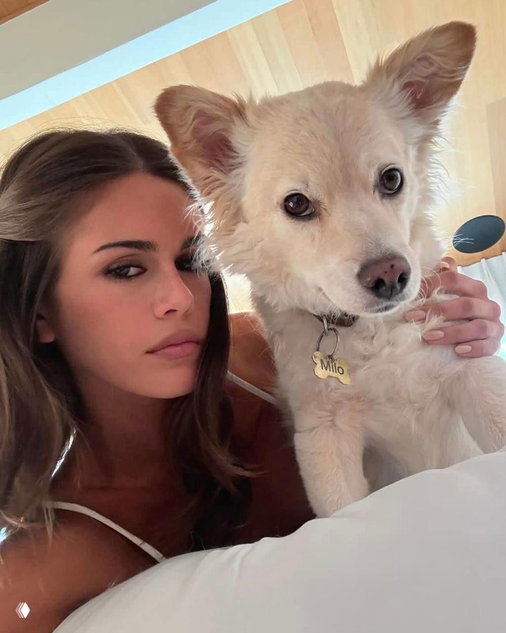 Крупный план молодая женщина рядом с кремовым пушистым псом; видно медальон с именем Milo, светлая спальня и спокойная атмосфера.