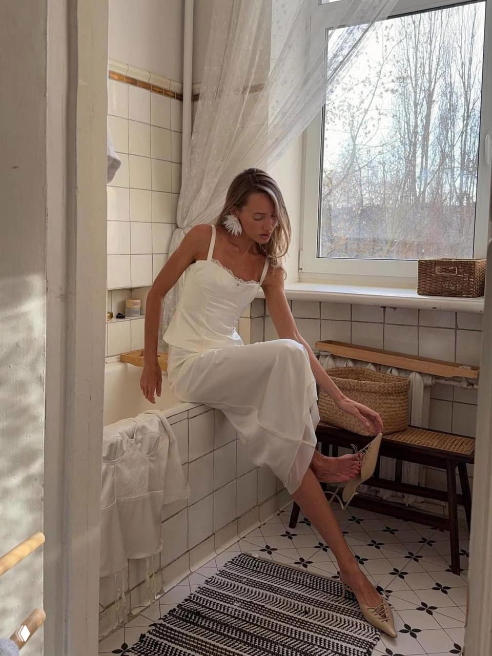 Женщина сидит на краю ванны в светлом платье, мягкий дневной свет через окно, атмосфера уюта и спокойствия.
