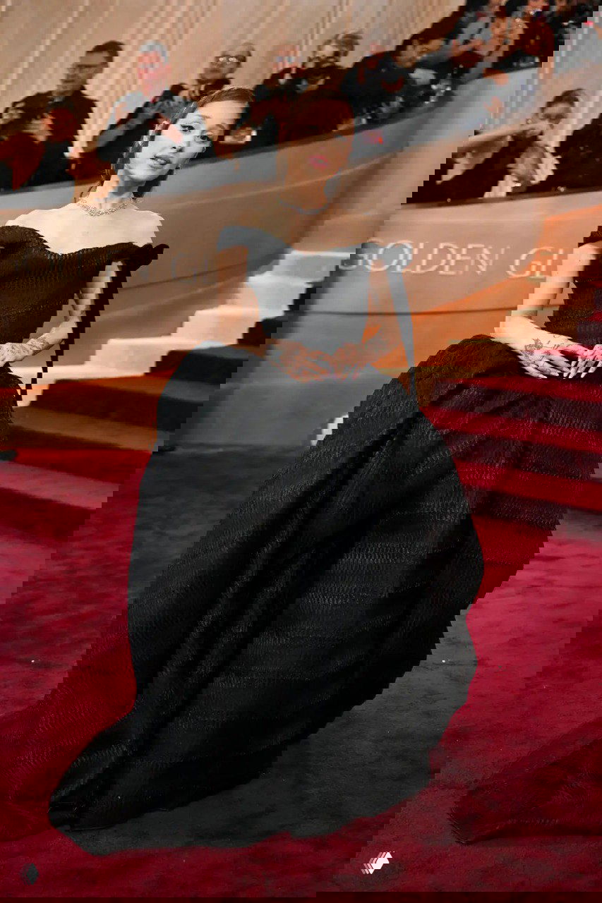 Женщина в пышном черном бальном платье с открытыми плечами и массивной юбкой стоит на красном ковре у сцены церемонии в окружении фотографов.