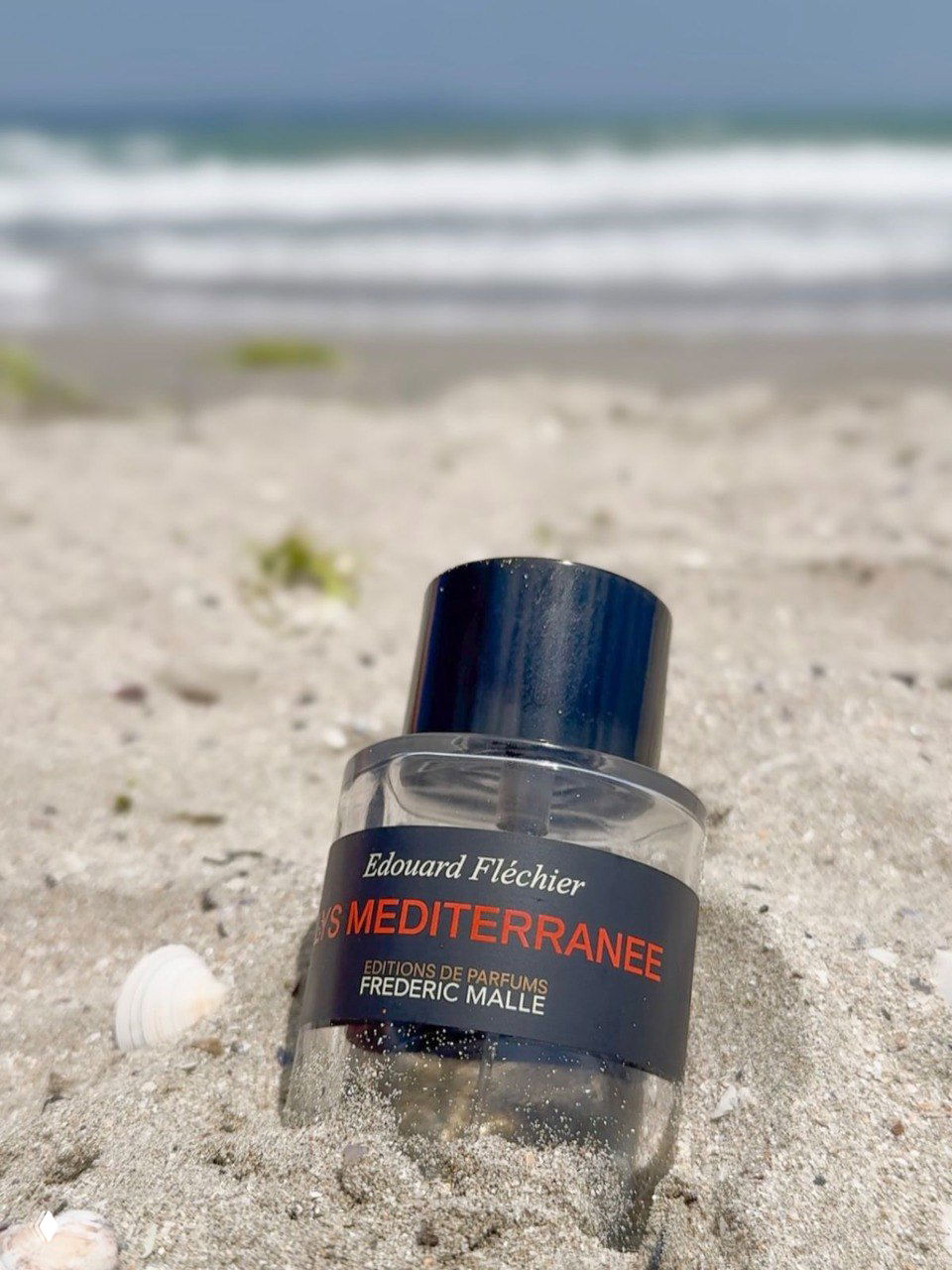 Флакон Frederic Malle Lys Mediterranee на песчаном берегу: бутылка частично в песке на фоне моря и ракушки, передающая морское и солнечное настроение аромата.
