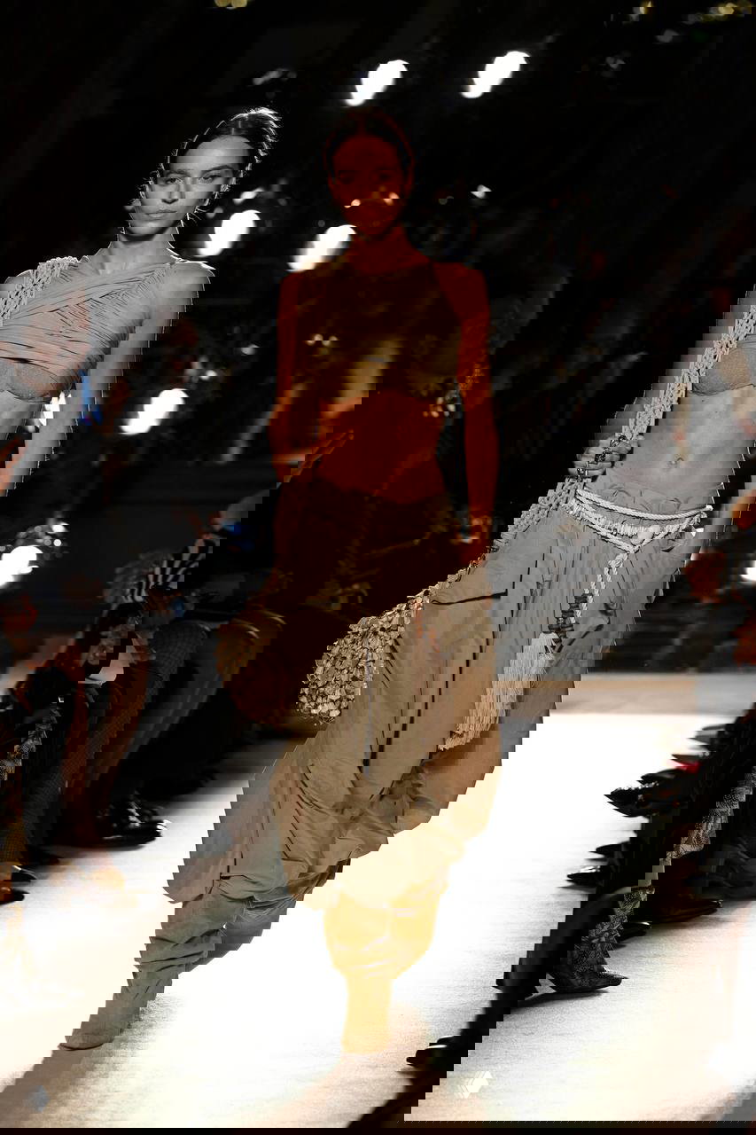 Модель в стиле бохо: топ‑бинго и широкие штаны с аксессуарами и плетёной сумкой под сценическим светом — богемная эстетика на подиуме.