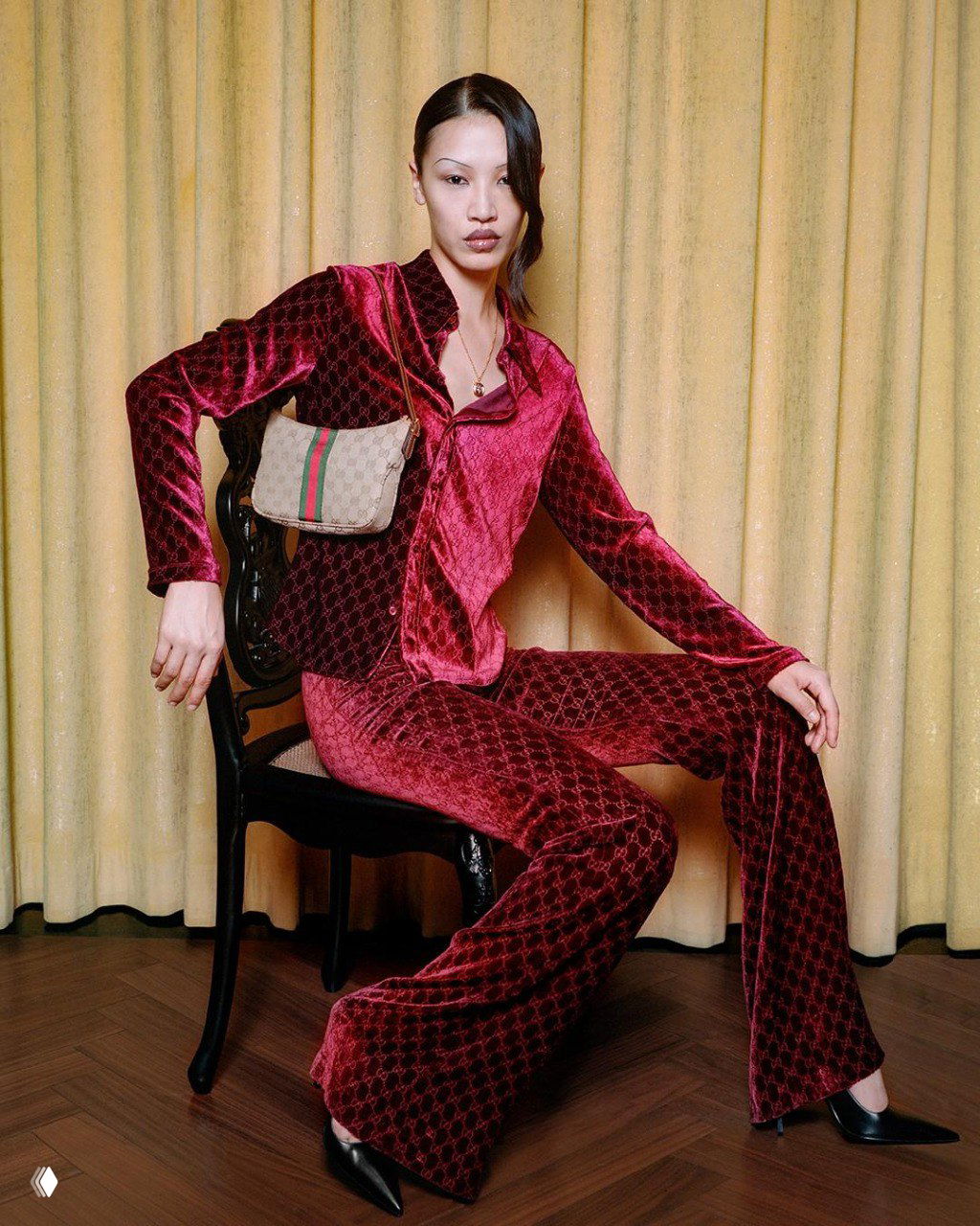 Женщина в бархатном бордовом костюме и сумкой Gucci сидит на стуле на фоне жёлтых штор — кадр из кампейна The Gathering.