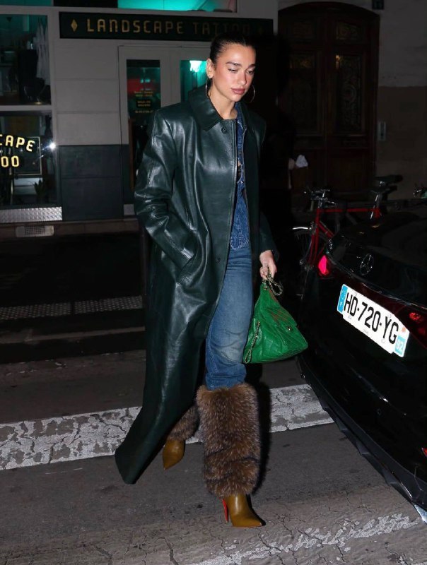 Девушка в длинном тёмно-зелёном кожаном пальто и джинсах выходит ночью на городской улице; вокруг припаркованные машины и фонари.