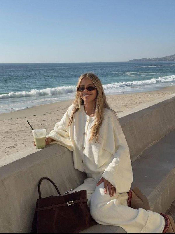 Женщина в светлом уютном костюме сидит у моря на набережной с сумкой рядом, мягкий дневной свет и морской фон.