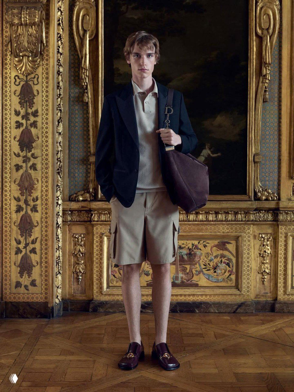 Модель в тёмном пиджаке и бежевых шортах держит сумку с металлической буквой D, сочетая спортивную рубашку-поло и традиционный декор дворянского образа в интерьере.