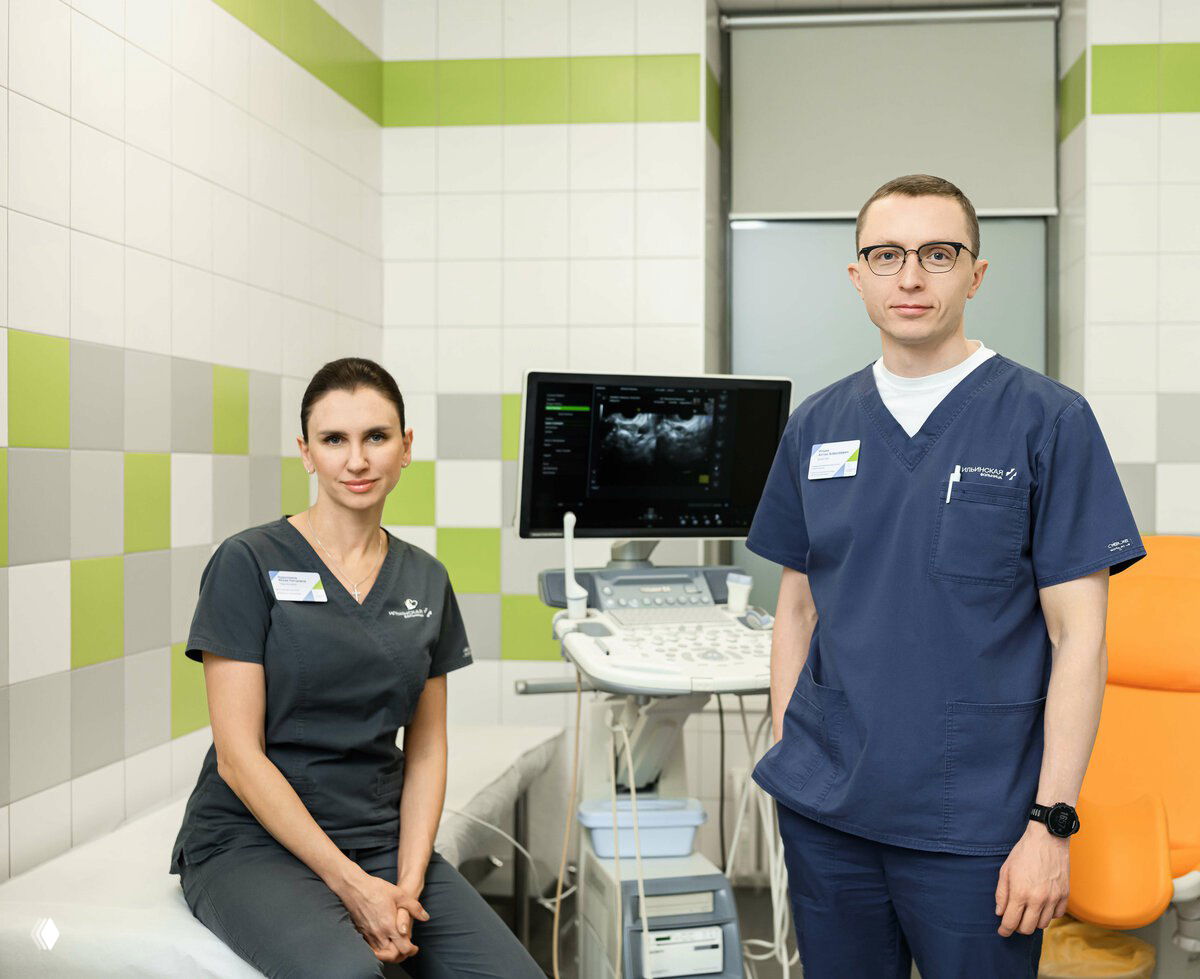 Двое медработников в кабинете с УЗИ-аппаратом: врач и специалист в медицинской форме на фоне диагностического оборудования.