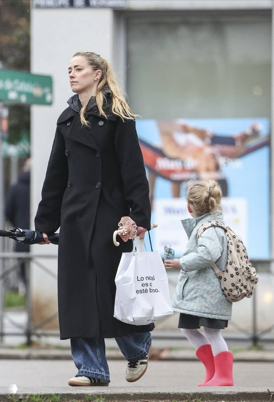 Женщина в тёмном пальто идёт по городскому тротуару, держит пакет и зонт; рядом идёт ребёнок в плаще и розовых резиновых сапожках.