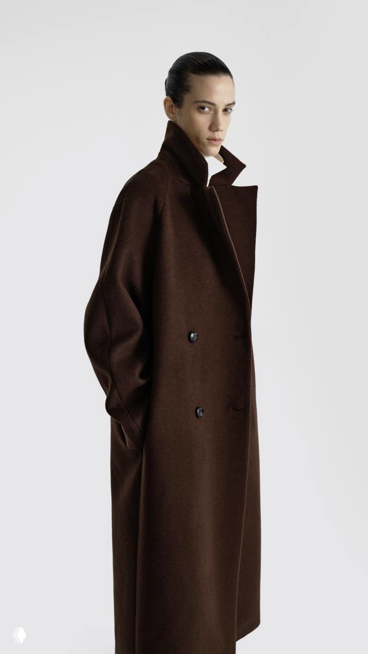 Модель в благородном коричневом двубортном пальто с поднятым воротником, акцент на чистой линии силуэта и плотной текстуре ткани на светлом фоне.