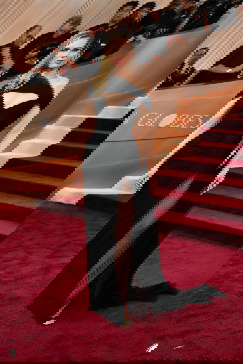 Женщина в черном платье с асимметричным вырезом и высоким разрезом позирует на красной дорожке у лестницы и фотозоны церемонии.