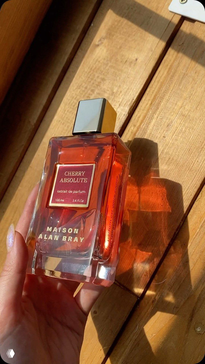 Флакон Alan Bray Maison Cherry Absolute в солнечном свете, который подчёркивает тёплые красные оттенки жидкости и отражение на деревянной поверхности.