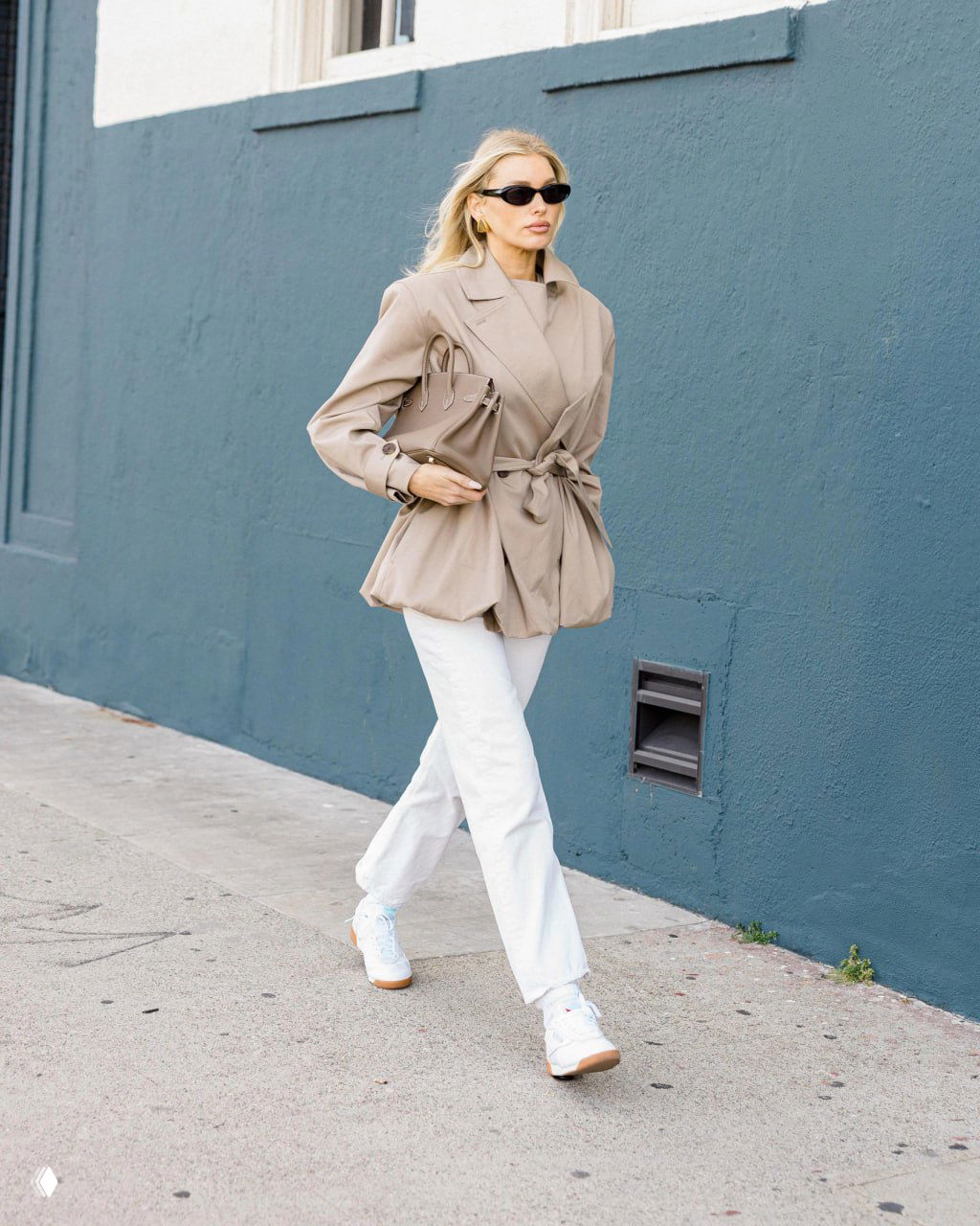 Женщина в бежевом жакете с поясом и белых брюках идет по тротуару в кроссовках, минималистичный городской фон, уличная съемка.
