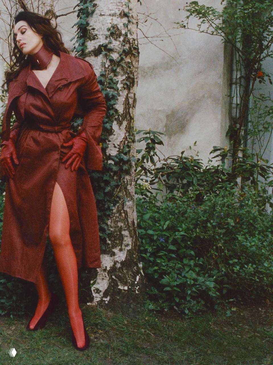 Уличная сцена: женщина в длинном плаще и красных перчатках позирует у берёзы и густой зелени, съёмка с атмосферным естественным светом.