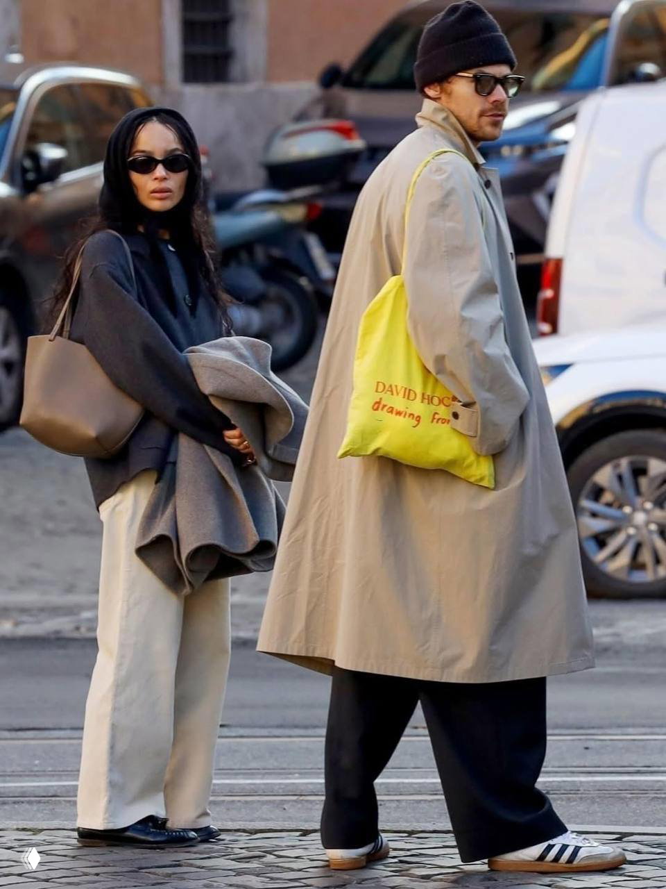 Зои и Гарри на улице в повседневных слоях: Зои в тёмном свитере и светлых брюках, Гарри в тренче и яркой сумке‑отоображении — образ в стиле улиц модных столиц.