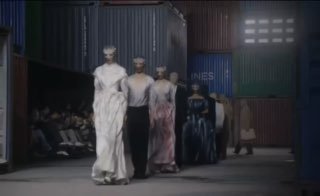 Кадр с подиума: несколько моделей идут по сцене из контейнеров в длинных платьях Maison Margiela, видна общая хореография и сценография.