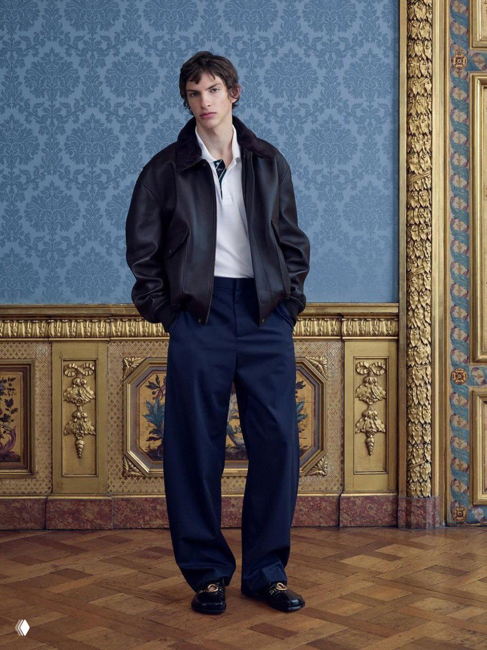 Модель в коричневой кожаной куртке и тёмно-синих брюках на фоне узорных обоев и золочёной рамы — акцент на материале и силуэте повседневного образа.