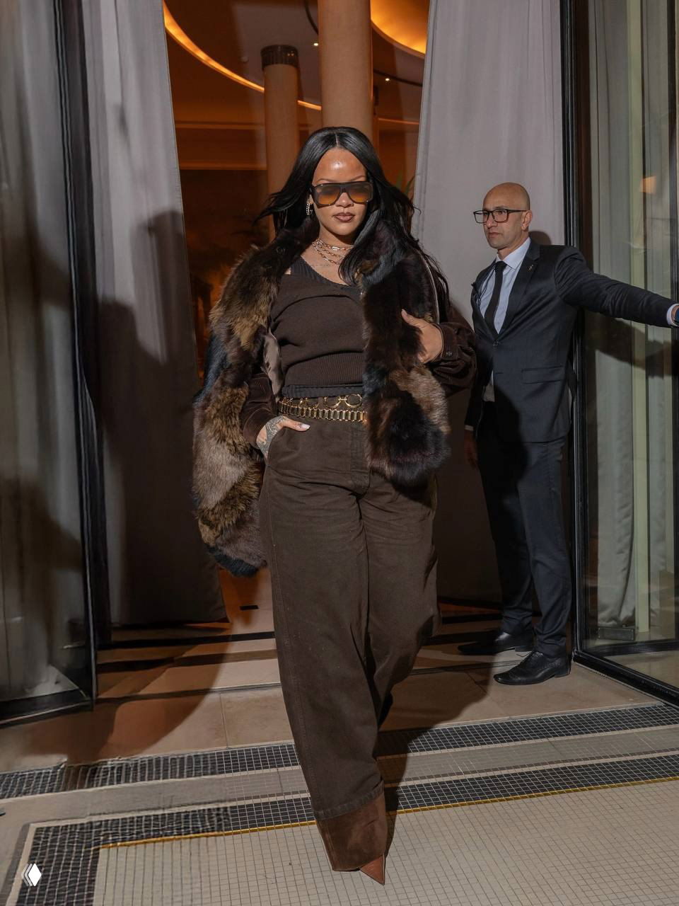 Женщина в коричневом образе с меховой накидкой и солнцезащитными очками выходит из здания; на заднем плане виден охранник и интерьер.