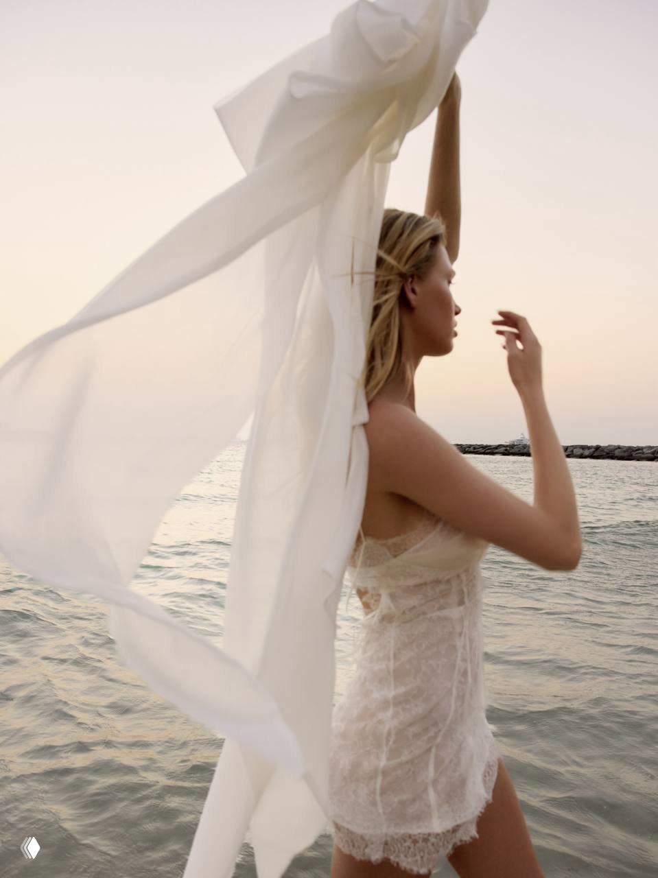 Профильная фотография модели у моря с развевающейся белой тканью: летняя редакционная композиция, легкое платье и аккуратный макияж.