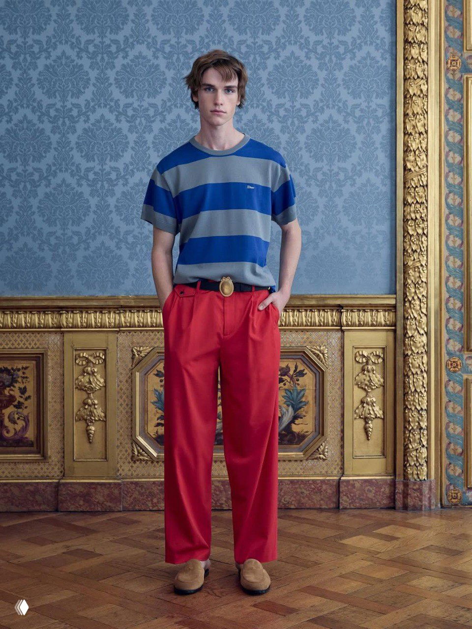 Молодой мужчина в синей полосатой футболке и ярко-красных брюках у декоративной панели, кадр подчёркивает контраст цвета и классический орнамент интерьера XVII века.