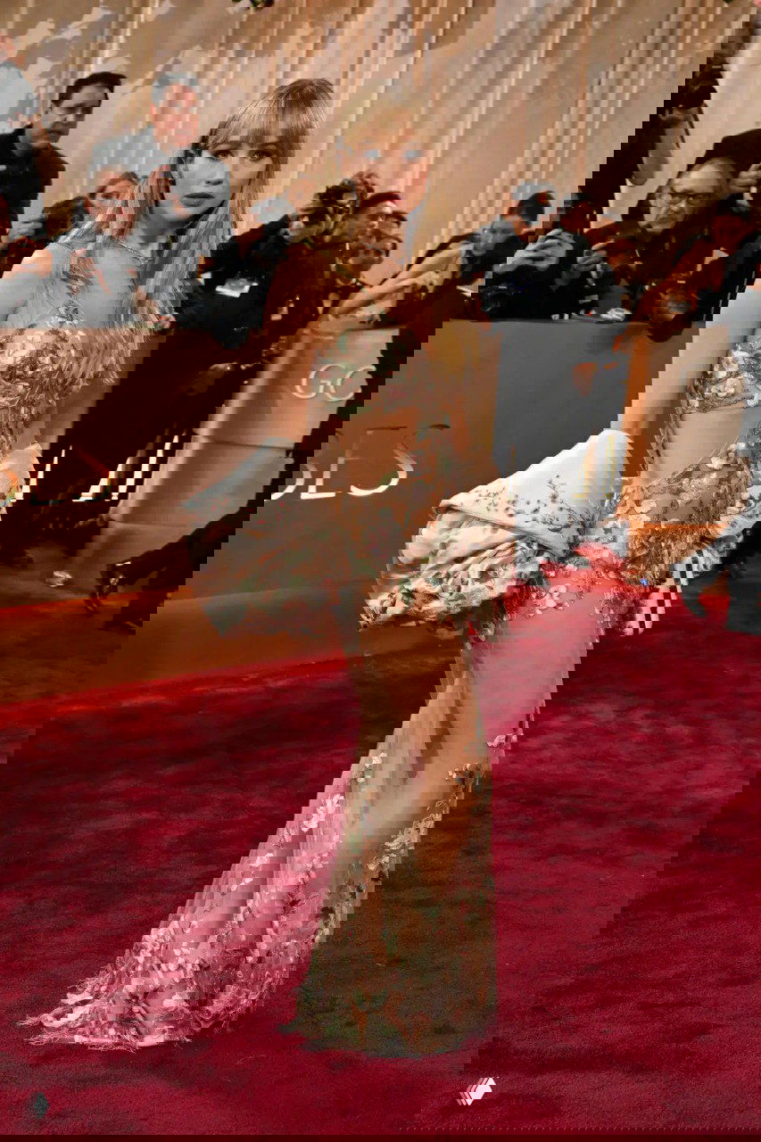 Женщина в полупрозрачном платье с цветочной вышивкой и оголенными участками позирует на красном ковре, вокруг — фотографы и зрители.