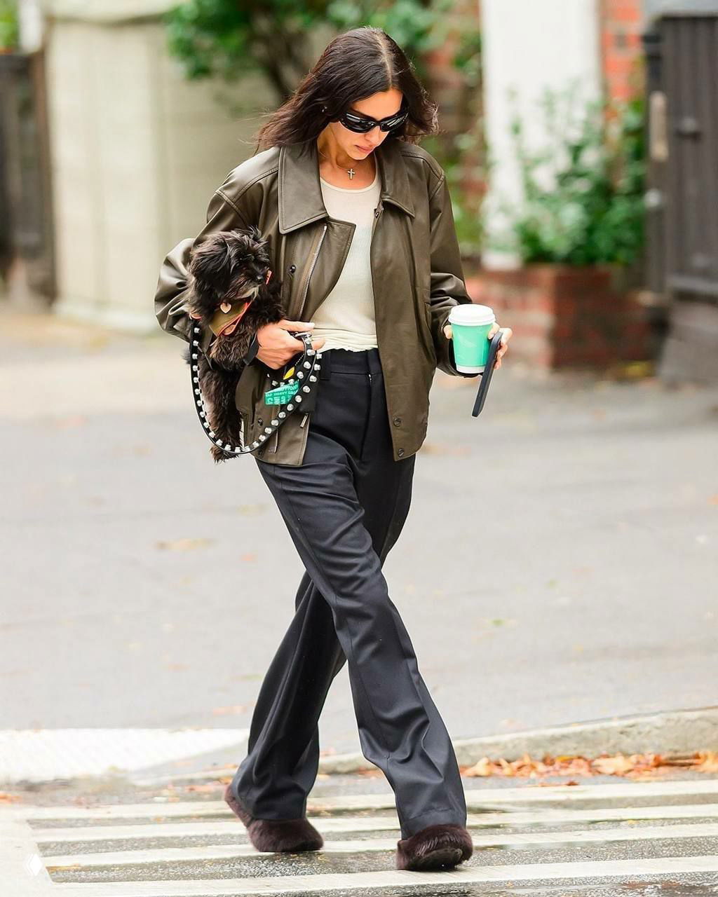 Папарацци: женщина в тёмной кожаной куртке и чёрных брюках держит маленькую собаку и бумажный стакан, уличная сцена.