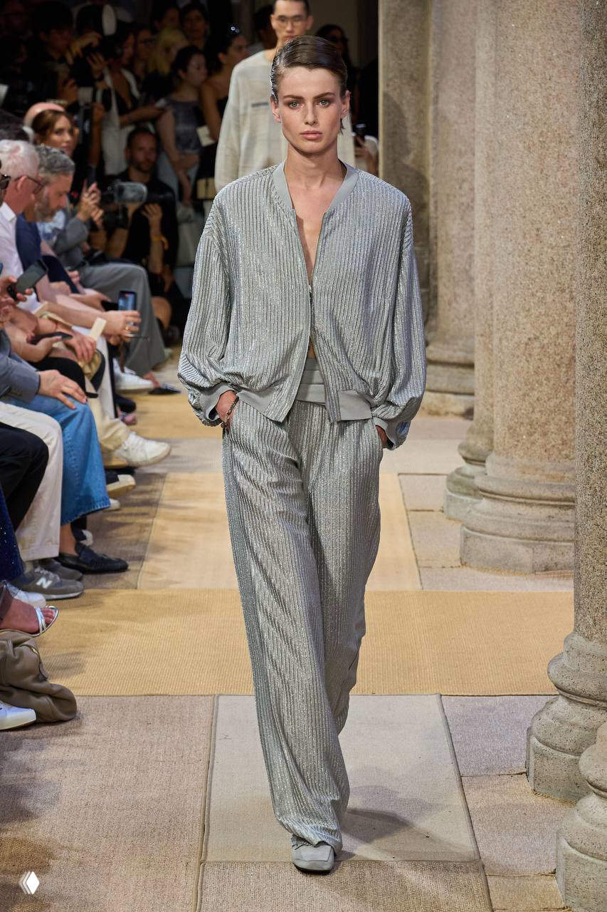 Женская модель в серебристом текстурном костюме идёт по дорожке показа Giorgio Armani в окружении зрителей и колонн исторического зала Милана.