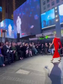 Женская модель в красном платье идёт по открытой площадке шоу в окружении зрителей и ночных рекламных экранов Таймс-сквер.