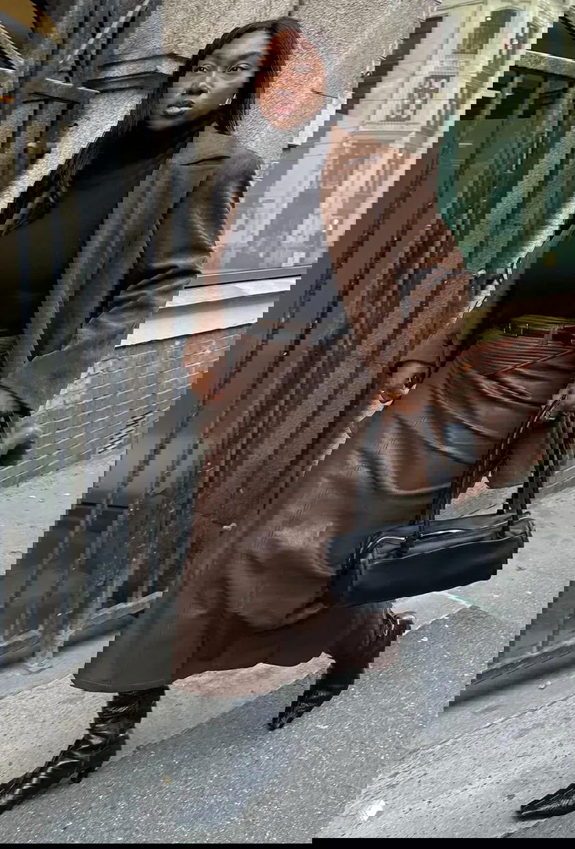 Женщина в чёрной водолазке, коричневом длинном кожаном пиджаке и юбке-миди, несущая чёрную сумку по городскому тротуару.