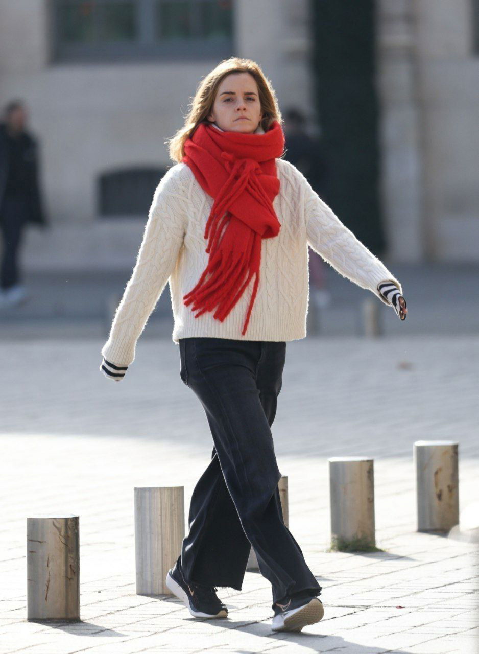 Уличный кадр: женщина в светлом вязаном свитере и ярком красном шарфе шагает по площади, фон — городской пейзаж.