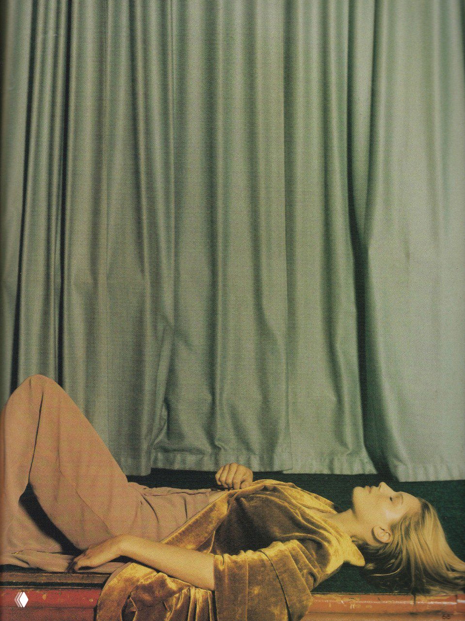 Никки Мохуд лежит на сцене или диване у закрытых штор, поза в расслабленном образе и бархатная текстура одежды в осенней палитре.