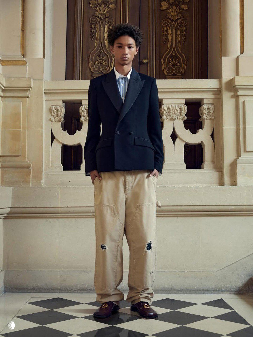 Модель в тёмно-синем двойном пиджаке и светлых мешковатых брюках на фоне светлой лестницы особняка — образ сочетает строгость кроя и свободный низ.